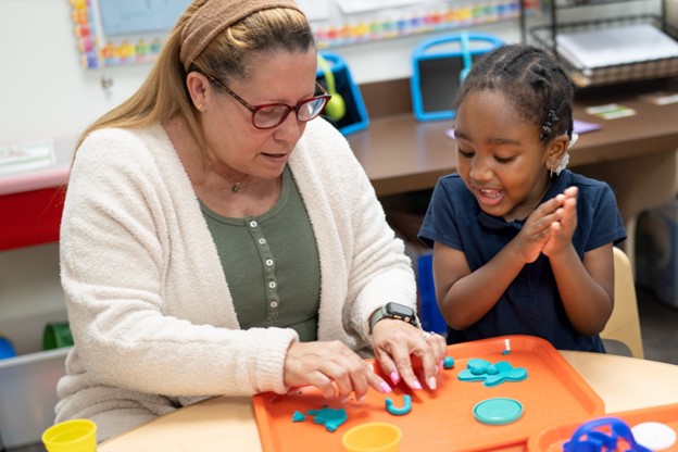 A preschool teacher sits with a pre-K student, playing with clay dough.
